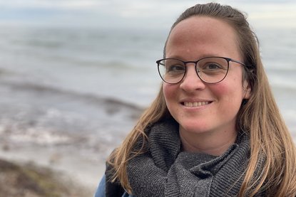 Eine Frau mit blonden langen Haaren und Brille schaut lächelnd in die Kamera. Im Hintergrund ist ein größeres Gewässer mit leichten Wellengang und ein Stück Strand zu sehen - Copyright: Maria Gutjahr