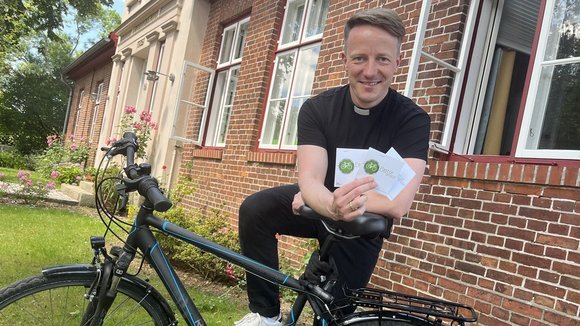 Ein junger Mann stützt sich auf ein Fahrrad, lächelt und hält zwei Karten in die Kamera.