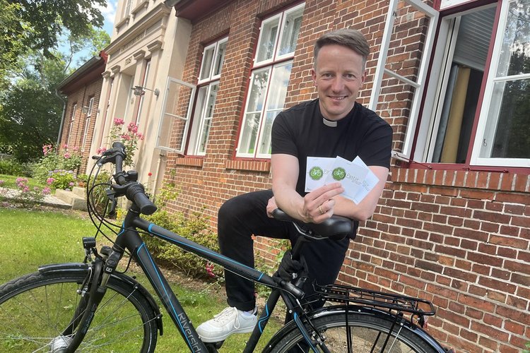 Ein junger Mann stützt sich auf ein Fahrrad, lächelt und hält zwei Karten in die Kamera. - Copyright: Annkathrin Bornholdt