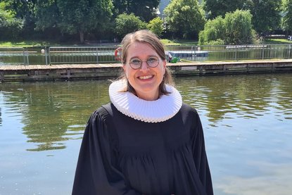 Eine Frau im Talar mit Brille schaut lächelnd in die Kamera. Im Hintergrund ist Wasser zu sehen und grüne Bäume und Sträucher - Copyright: Margrit Wegner