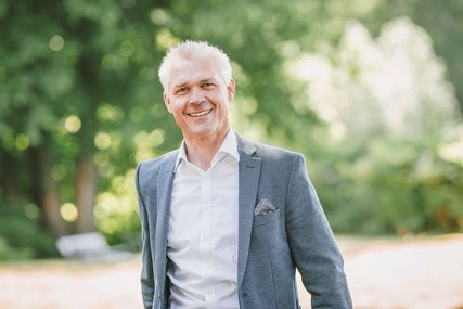 Ein Mann mit grauen kurzen Haaren trägt ein weißes Hemd und ein graues Jackett. Im Hintergrund befinden sich etwas verschwommen grüne Bäume und Sträucher. - Copyright: Guido Kollmeier