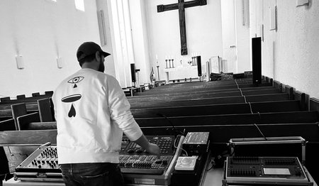 Ein Mann von hinten zu sehen in einem Kirchenraum mit Kreuz und DJ Technik.