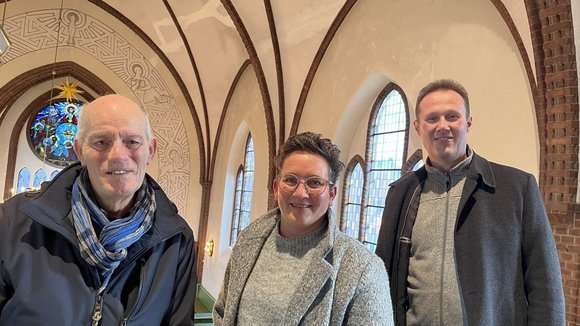 Zwei Männer und eine Frau stehen auf der Empore einer Kirche mit dem Gewölbe im Hintergrund.