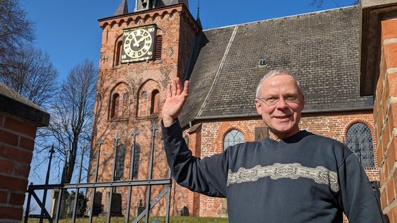 Ein fröhlicher Mann schaut winkend in die Kamera.