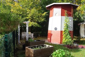 Ein breiter Leuchtturm aus Holz steht in einem Garten umgeben von Hochbeeten.