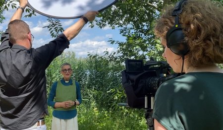 Pastorin Katja von Kiedrowski wird in einer üppigen Naturlandschaft gefilmt. Eine Kamera zeigt in ihre Richtung.