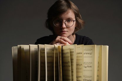 Ein offenes Notenbuch, aufgefächert steht hochkannt, eine Frau mit Brille und kurzen braunen Haaren hat ihre Hände übereinander aufgestützt, ihr Kinn auf diesen Händen und schaut in die Kamera - Copyright: Nataliya Abryutina