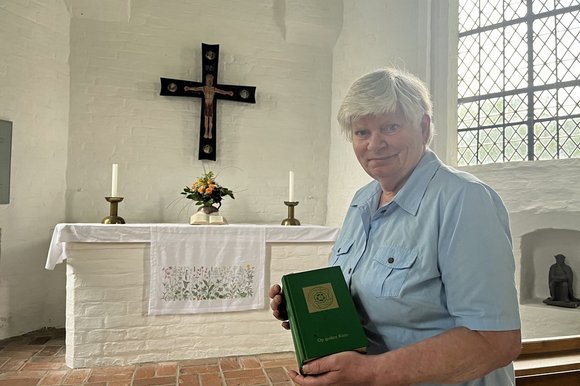 Eine Frau mit kurzen grauen Haaren steht vor einem Altar und hält ein Liederbuch in der Hand.