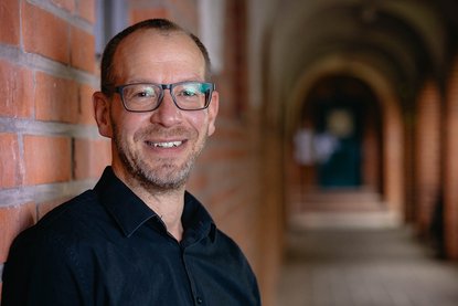 Ein Mann lächelt mit sehr kurzen Haaren und Brille in die Kamera. Er lehnt an einer roten Ziegelwand und rechts sieht man verschwommen einen altertümlichen Gang. - Copyright: Mathias Hollaender