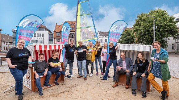 Mehrere Menschen auf einer Sandfläche mit Strandkörben lächeln in die Kamera.