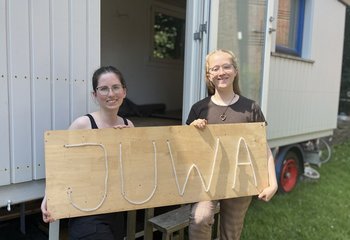 Zwei junge Frauen stehen vor einem Bauwagen und halten ein Schild mit der Aufschrift 'JuWa'. - Copyright: Annkathrin Bornholdt