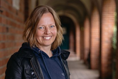 Eine Frau mit mittellangen blonden Haaren lächelt in die Kamera. Sie ist dunkel gekleidet. Hinter ihr sieht man einen altertümlichen Gang und links eine rote Backsteinmauer - Copyright: Mathias Hollaender