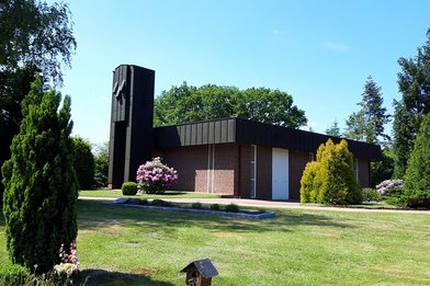 Kapelle Waldfriedhof steht im Hintergrund bei strahlend blauem Himmel. Links und rechts sind Bäume und Sträucher. Im Vordergrund Rasen und links ein kleiner grüner Baum - Copyright: Ev.-Luth. Kirchengemeinde Büchen-Pötrau